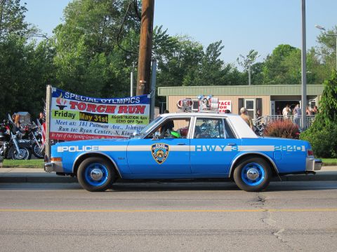 1991 Ford LTD Crown Victoria - light-blue marked Police 'HWY' cruiser with a roof-mounted Special Olympics Torch Run banner, parked along a street at an outdoor event.