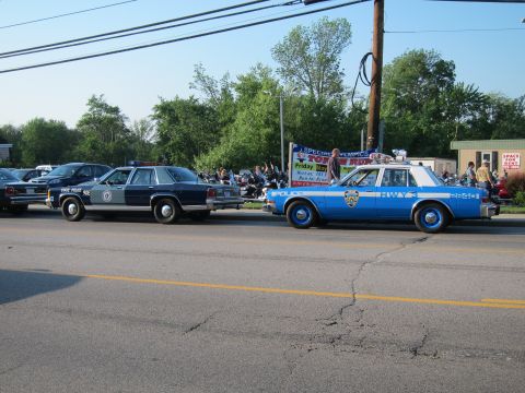 1991 Ford LTD Crown Victoria - light-blue marked Police 'HWY' cruiser parked behind a dark-blue State Police sedan along a cracked asphalt street with a crowd behind.