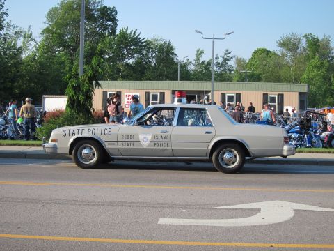 1991 Ford LTD Crown Victoria - silver Rhode Island State Police sedan sits curbside marked STATE POLICE as motorcycles and a crowd gather behind.