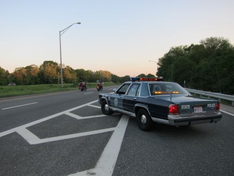 1991 Ford LTD Crown Victoria - dark State Police sedan with light bar parked across a highway lane at dusk as motorcycles approach.