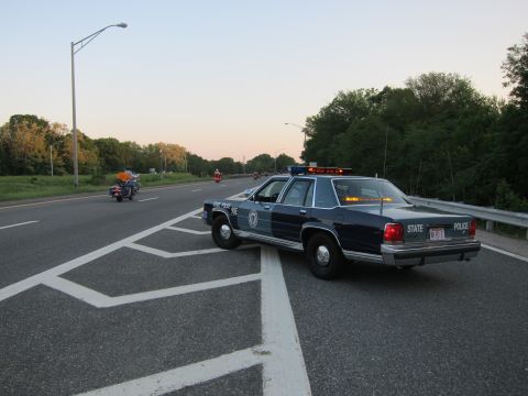 1991 Ford LTD Crown Victoria - dark State Police sedan with activated lights parked on a highway at dusk, motorcyclists in the distance.