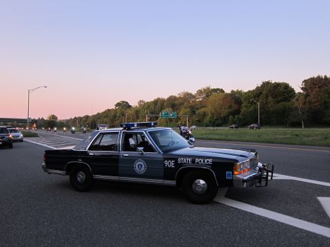 1991 Ford LTD Crown Victoria - dark Massachusetts State Police sedan marked 90E with front push bar on a highway at sunset.