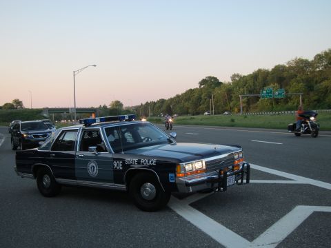 1991 Ford LTD Crown Victoria - dark State Police sedan marked 90E with front push bar sits on a highway lane at dusk near motorcycles.