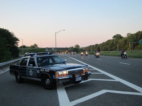 1991 Ford LTD Crown Victoria - dark Ford State Police sedan with push bar parked on a highway at dusk as a line of motorcycles approaches.