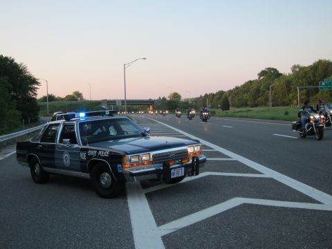 1991 Ford LTD Crown Victoria - dark State Police sedan marked 90E with light bar parked on a highway at sunset, motorcycles riding past.