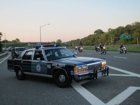 1991 Ford LTD Crown Victoria - dark Ford State Police sedan marked 90E with front push bar on a highway at dusk as motorcycles pass by.