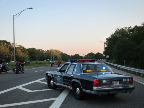 1991 Ford LTD Crown Victoria - Rear view of a dark State Police sedan marked 90 E parked on a highway at dusk as motorcycles ride past.