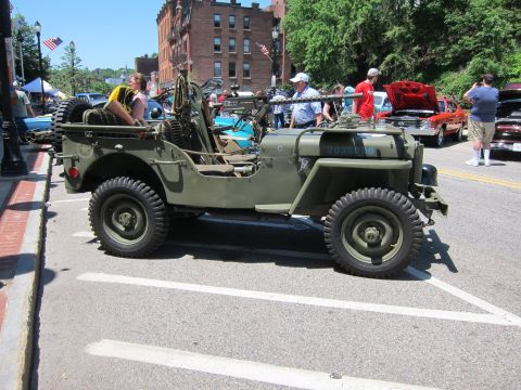 1991 Ford LTD Crown Victoria - olive drab military jeep with a mounted rifle parked on a downtown street at a car show with brick buildings.