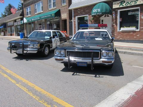 1991 Ford LTD Crown Victoria - Two vintage black police cars, one CHP-marked, parked nose-out on a sunny street outside a barber shop.