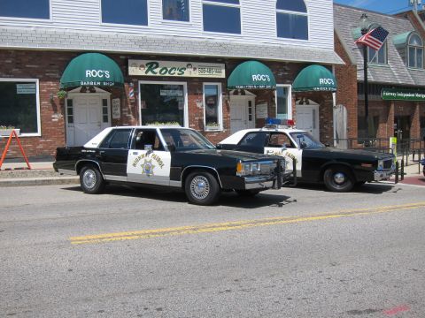 1991 Ford LTD Crown Victoria - Two black-and-white Highway Patrol sedans parked side by side in front of Roc's barber shop.