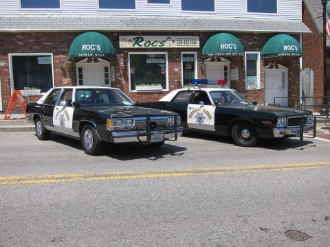1991 Ford LTD Crown Victoria - modern-era Highway Patrol sedan beside an older black-and-white Highway Patrol car in front of Roc's storefronts.