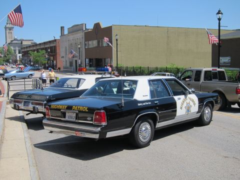 1991 Ford LTD Crown Victoria - Rear three-quarter view of a black-and-white Highway Patrol sedan parked beside a tan SUV near brick buildings and flags.