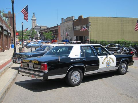 1991 Ford LTD Crown Victoria - Rear three-quarter view of a black-and-white Highway Patrol sedan parked at a downtown curb with a tower in the background.