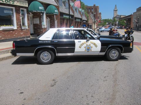 1991 Ford LTD Crown Victoria - Side profile of a black-and-white Highway Patrol sedan with motorcycles parked behind it on a downtown street.
