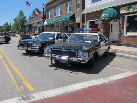 1991 Ford LTD Crown Victoria - Two vintage black-and-white police sedans angle-parked along a downtown street with US flags and storefronts behind.