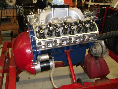 1970 Ford Mustang - A V8 engine is assembled on a red stand, with a red flywheel housing and blue oil pan visible.