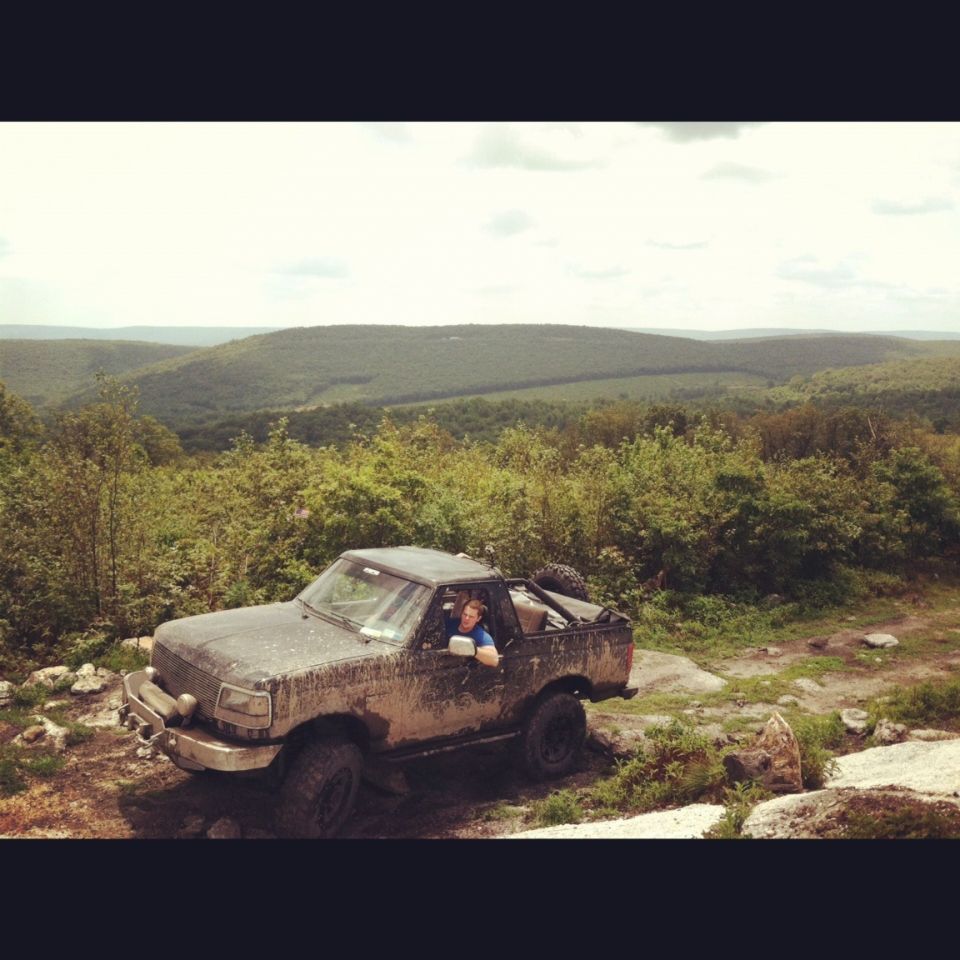 Muddy 1990s Ford Bronco parked on a rocky, wooded incline with a person inside