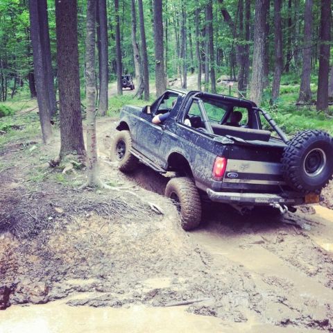 1995 Ford Bronco - A 1995 Ford Bronco tackles a challenging muddy path in a wooded area, with another vehicle visible in the distance.