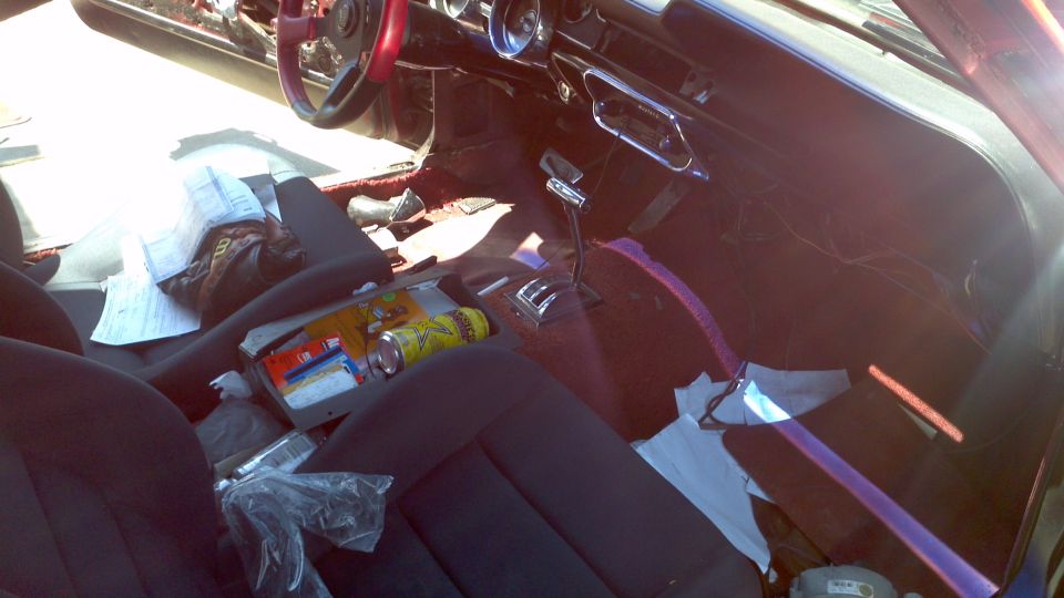 Car interior with dark seats, red steering wheel, and a chrome gear shifter. Papers and cans are scattered on the seats and floor