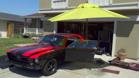 1987 Ford Bronco - Black 1967 Ford Mustang with red racing stripes parked in front of a garage, driver door open.