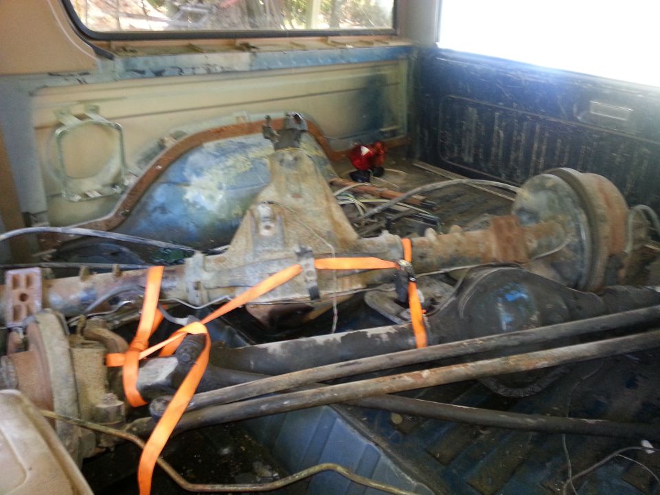 Rusty rear axle with differential strapped with orange strap inside a worn blue truck cargo bed