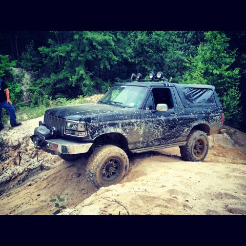 1995 Ford Bronco - Mud-covered 1995 Ford Bronco navigates a sandy, off-road trail with aftermarket lights and winch.