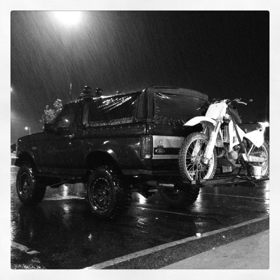 Black Ford Bronco with a dirt bike mounted on the rear rack, parked on a wet street at night