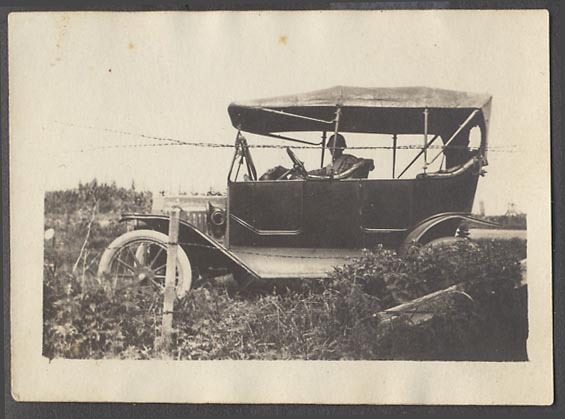 Side view of a touring car with a soft top, driver and passenger visible, parked near a barbed wire fence