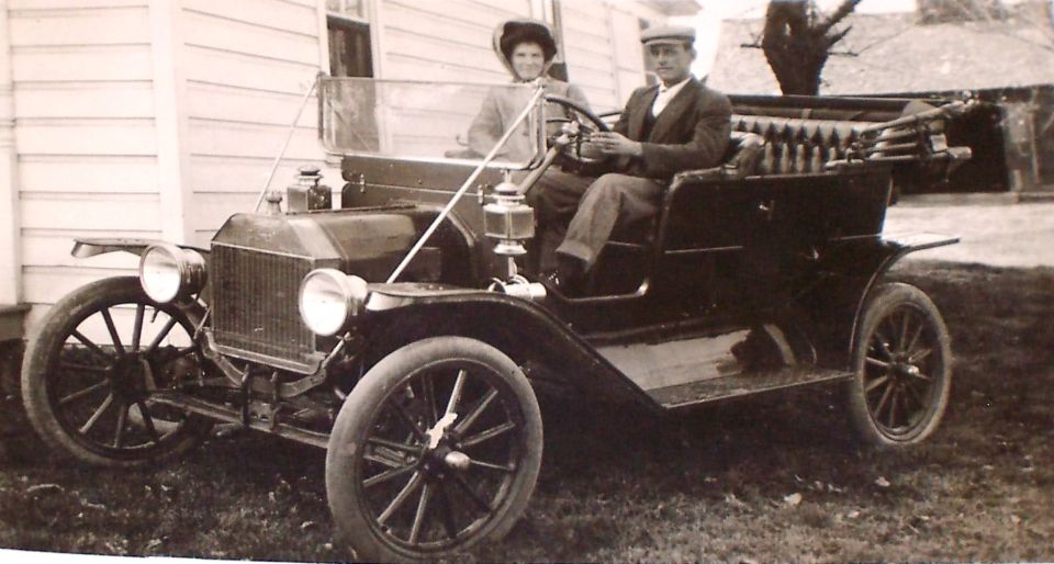 Antique photo of a couple seated in a brass-era Ford Model T touring car by a clapboard house