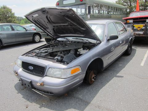 1991 Ford LTD Crown Victoria - A gray Ford Crown Victoria sits with its hood open and one wheel missing, parked next to other vehicles.