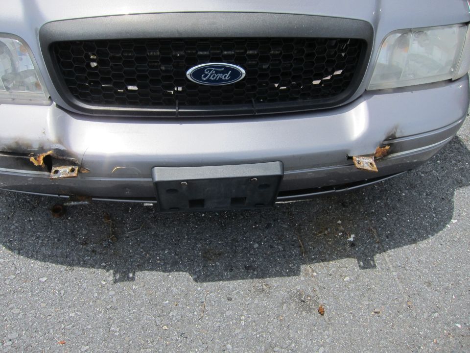 Front of a silver Ford Crown Victoria with a black grille and a Ford logo. The bumper is damaged and rusted on both sides