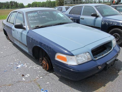1991 Ford LTD Crown Victoria - A damaged police car sits in a lot with other vehicles, missing its wheels and front grille.