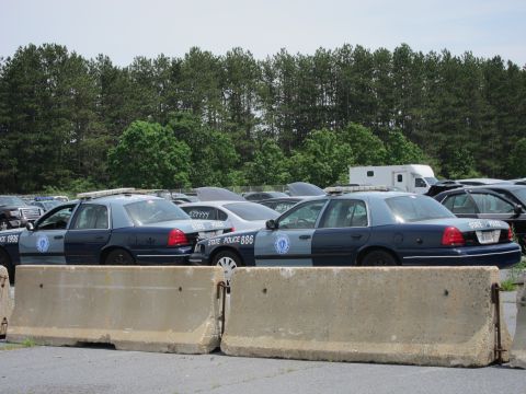 1991 Ford LTD Crown Victoria - A line of state police vehicles are parked in an outdoor lot, with concrete barriers in the foreground.