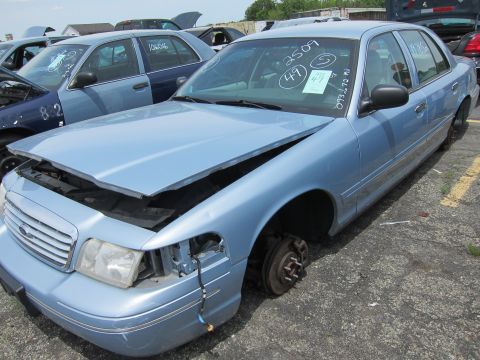 1991 Ford LTD Crown Victoria - A light blue sedan sits in a junkyard with its hood ajar and one wheel missing.