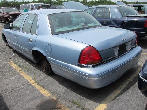 1991 Ford LTD Crown Victoria - A light blue sedan, missing its wheels, sits in a junkyard. The body style suggests a 2003 model year.