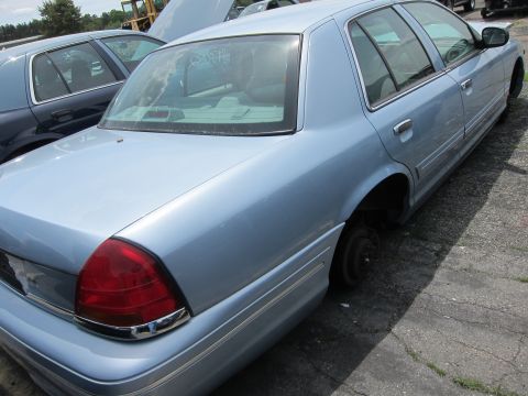 1991 Ford LTD Crown Victoria - Rear view of a light blue sedan, likely a 1998-2002 Ford Crown Victoria, at a junkyard.