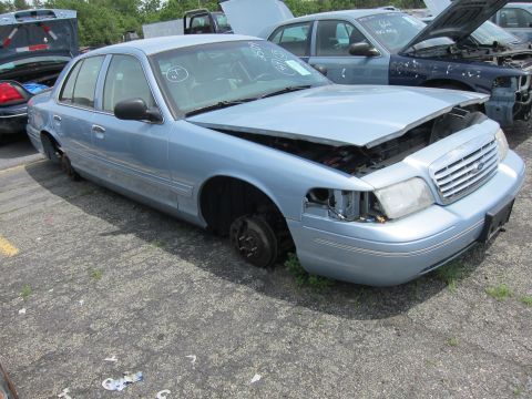 1991 Ford LTD Crown Victoria - A light blue sedan sits in a junkyard with its wheels removed and hood ajar, awaiting parts or disposal.