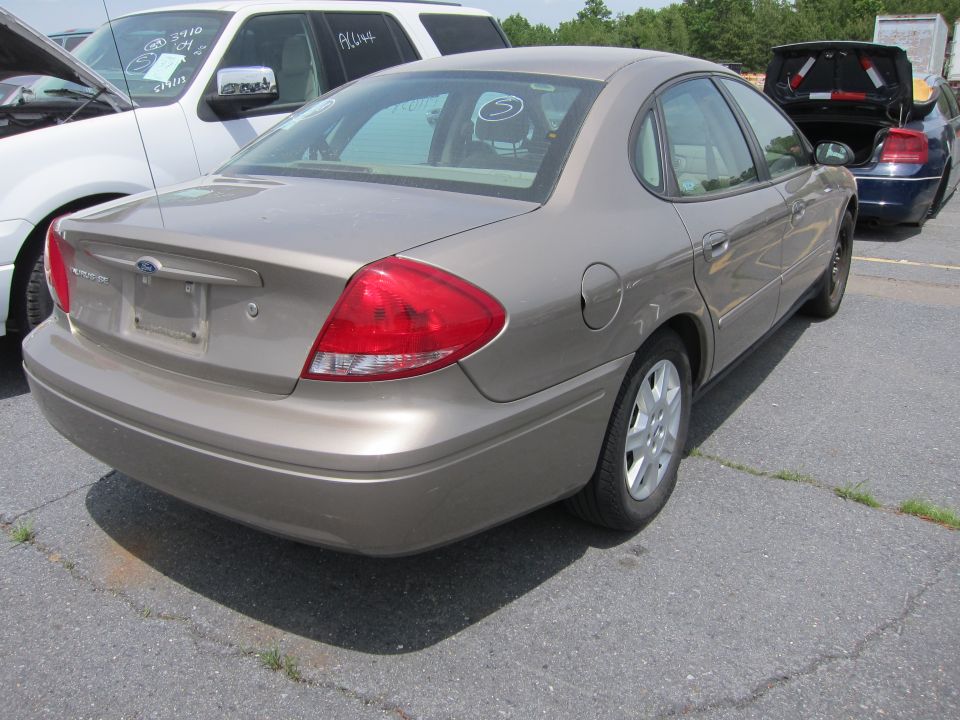 Tan Ford Taurus sedan parked on asphalt, rear view. White SUV and blue car in background