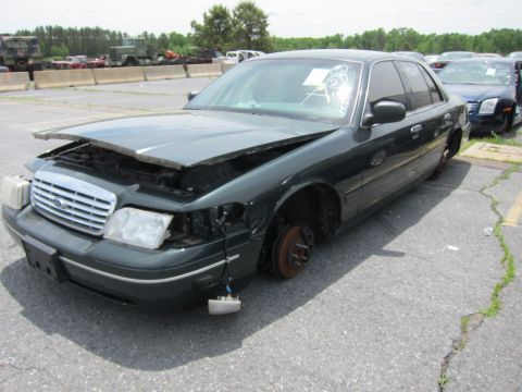 1991 Ford LTD Crown Victoria - A damaged dark green Ford Crown Victoria sits in a lot, missing a wheel and headlight, with its hood ajar.