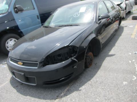 1991 Ford LTD Crown Victoria - A damaged black sedan sits in a lot next to a blue van, missing its front passenger wheel and headlight.