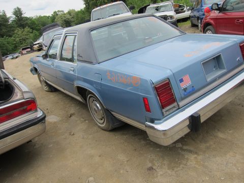 2001 Ford Crown Victoria - A blue sedan with a black roof sits in a junkyard, showing orange graffiti and an American flag sticker on its trunk.
