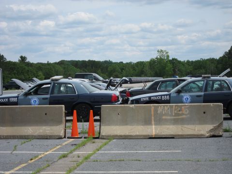 1991 Ford LTD Crown Victoria - Multiple police vehicles are visible behind concrete barriers under a cloudy sky.
