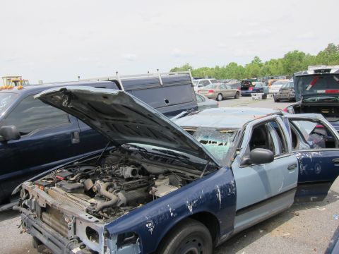 1991 Ford LTD Crown Victoria - A damaged sedan sits in a junkyard with its hood and doors open, revealing the engine compartment.