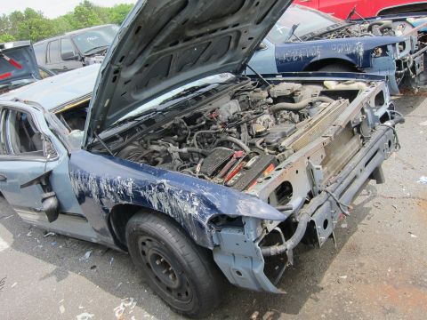1991 Ford LTD Crown Victoria - A heavily damaged blue sedan sits in a junkyard with its hood open, showing the engine compartment.