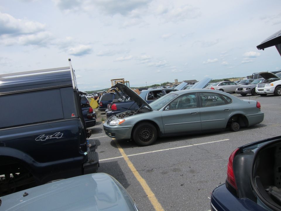 Gray sedan in junkyard, hood up, missing front wheel. Dark blue pickup truck to the left