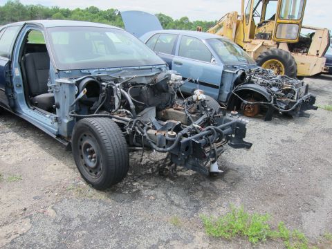 1991 Ford LTD Crown Victoria - Two heavily stripped blue sedans sit in a junkyard, one with its hood open, next to a yellow piece of heavy machinery.