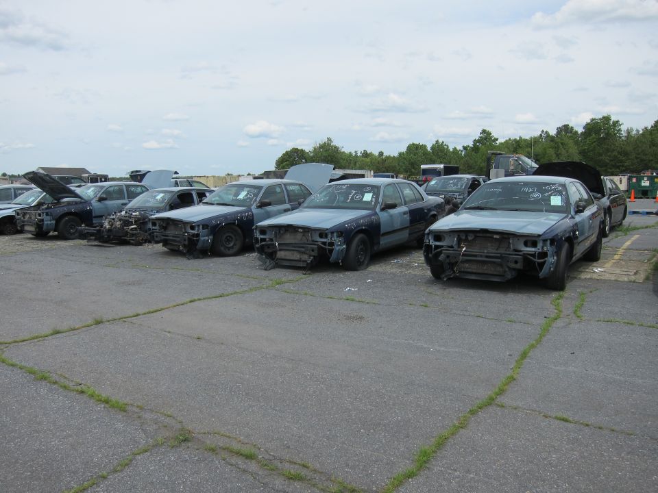 Several Ford Crown Victorias in a junkyard, front ends missing. Some have handwritten tags on windshields