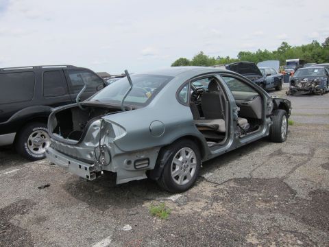 1991 Ford LTD Crown Victoria - A stripped-down gray sedan sits in a junkyard, missing its doors and trunk, with other vehicles visible in the background.