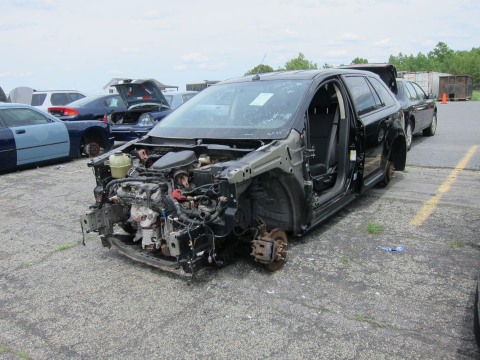 Front end of a black SUV is missing, showing engine components. Wheels are gone. Other cars in background