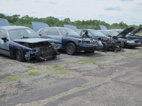 1991 Ford LTD Crown Victoria - Several damaged police cruisers are parked in a lot, some with hoods open, awaiting repair or dismantling.